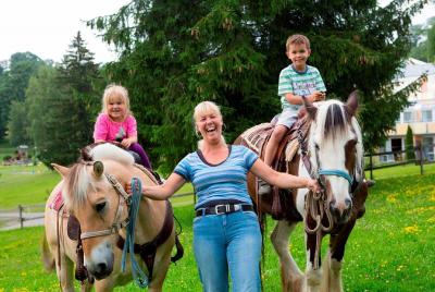 Zwei Kinder reiten auf Ponys, zwischen ihnen lacht eine Frau in die Kamera